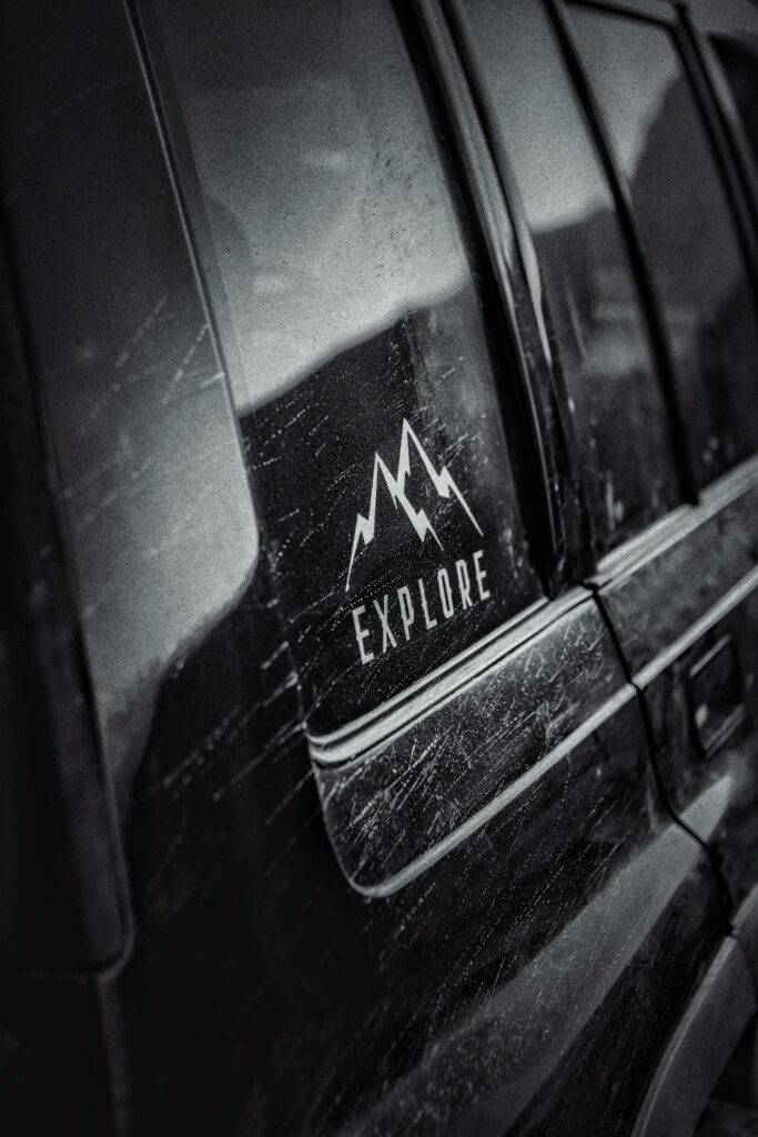 Close-up of a dusty car window featuring an Explore sticker, captured in black and white.